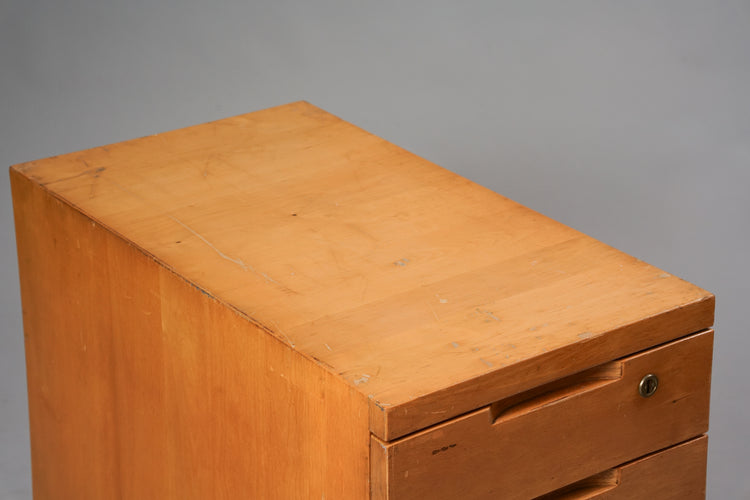 Wooden filing cabinet with five drawers. The cabinet has a lock on the first drawer and four wheels. Worn from use.
